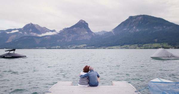 Rear view of senior pensioner couple tourists sitting on pier by lake, hugging.