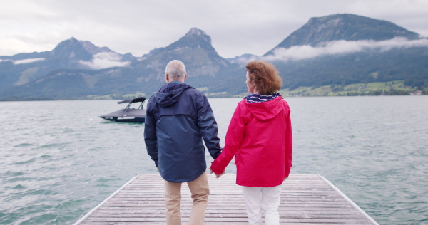 Rear view of senior pensioner couple tourists walking on pier by lake, holding hands.