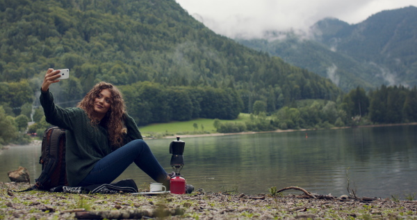 Young woman with curly hair tourist on a hiking trip in nature, sitting and taking selfie.