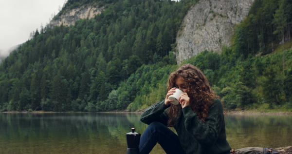 Young woman tourist with curly hair on a hiking trip in nature, resting, drinking coffee.