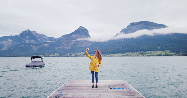 Portrait of young woman tourist standing on a pier by lake, taking selfie with smartphone.