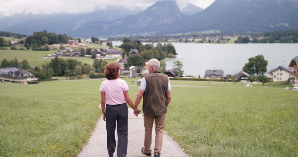 A rear view of active senior pensioner couple hiking in nature, holding hands.