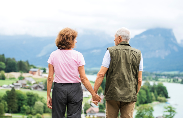 A rear view of senior pensioner couple hiking in nature, holding hands.