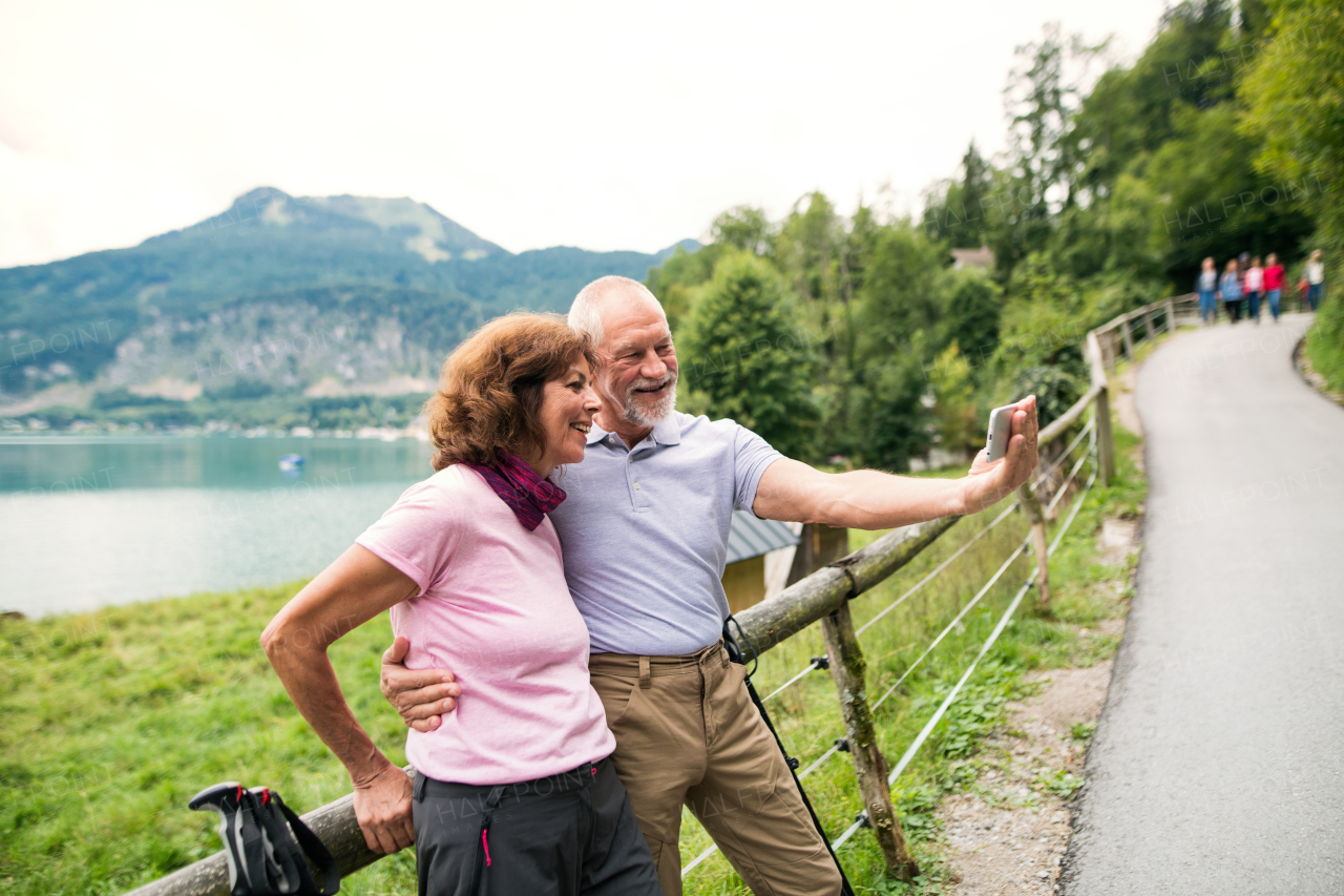 A senior pensioner couple with smartphone hikers standing in nature, taking selfie.