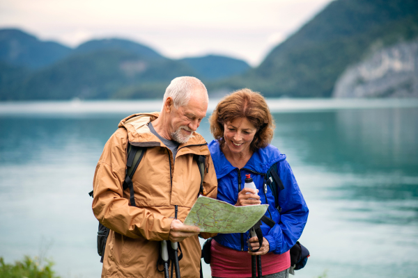 A front view of senior pensioner couple hikers standing by lake in nature, using map.