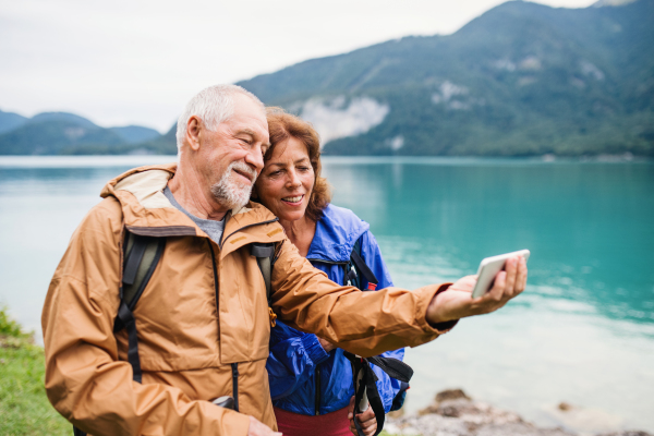 A senior pensioner couple with smartphne standing by lake in nature, taking selfie.