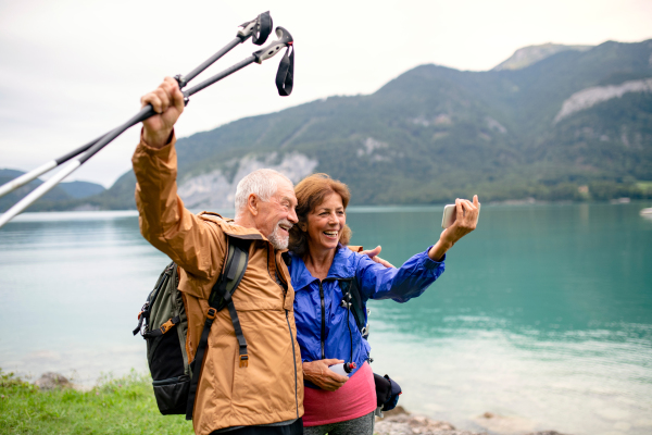 A senior pensioner couple with smartphne standing by lake in nature, taking selfie.