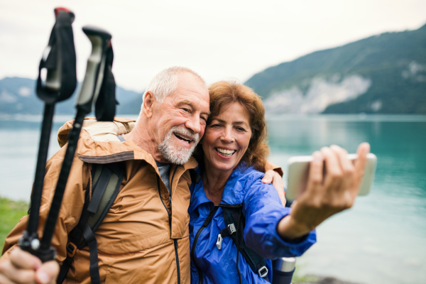 A senior pensioner couple with smartphne standing by lake in nature, taking selfie.