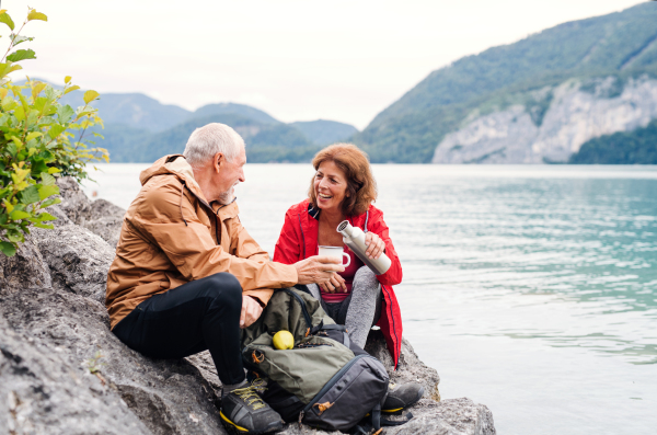 A cheerful senior pensioner couple hiking by lake in nature, sitting and resting.