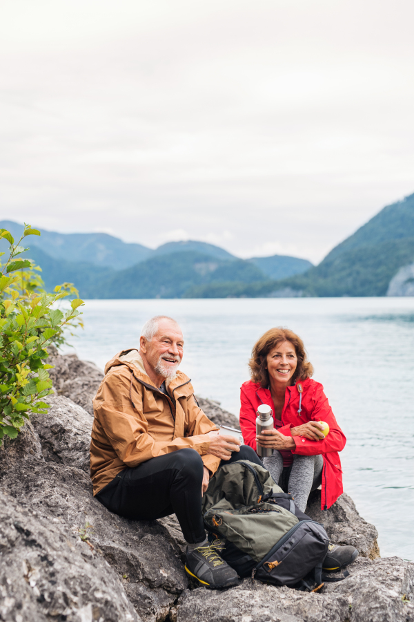 A cheerful senior pensioner couple hiking by lake in nature, sitting and resting.