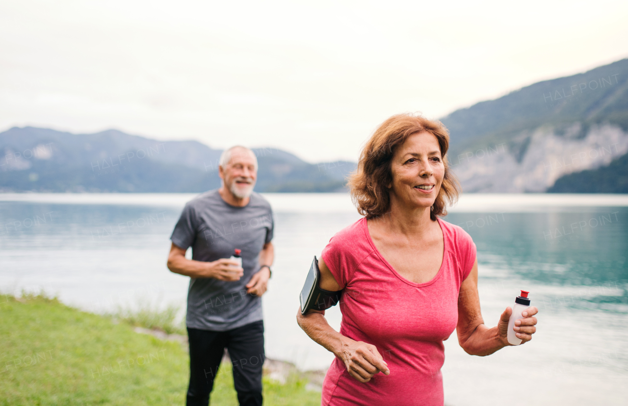 A senior pensioner couple with smartphone running by lake in nature.