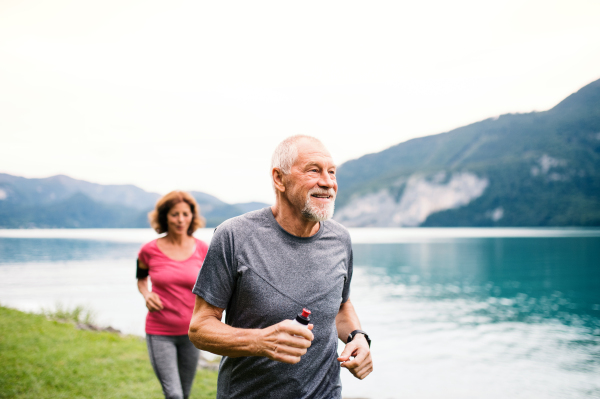 A senior pensioner couple with smartphone running by lake in nature.