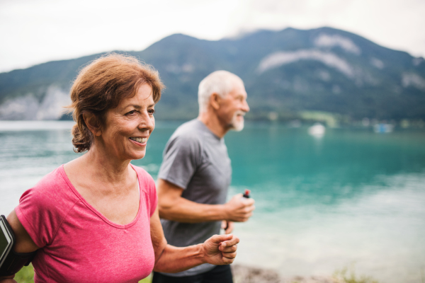 A senior pensioner couple with smartphone running by lake in nature.