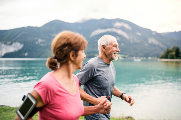 A senior pensioner couple with smartphone running by lake in nature.