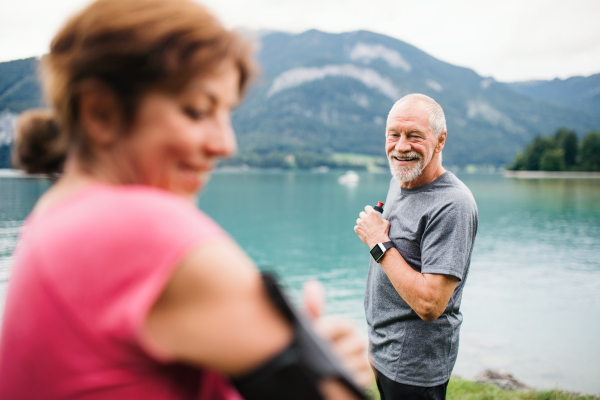 Senior pensioner couple runners with smartphone by lake in nature, resting.