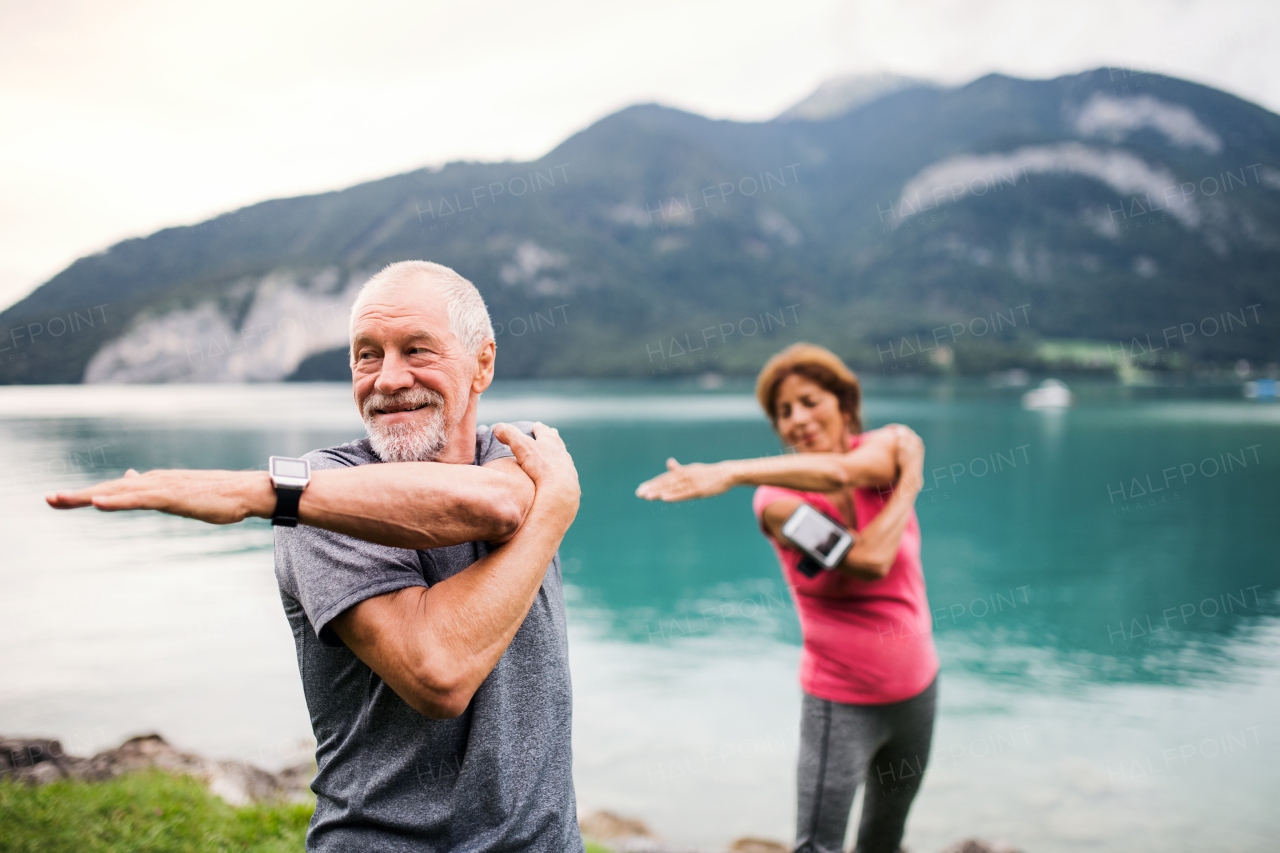 A senior pensioner couple with smartphone by lake in nature, doing exercise.