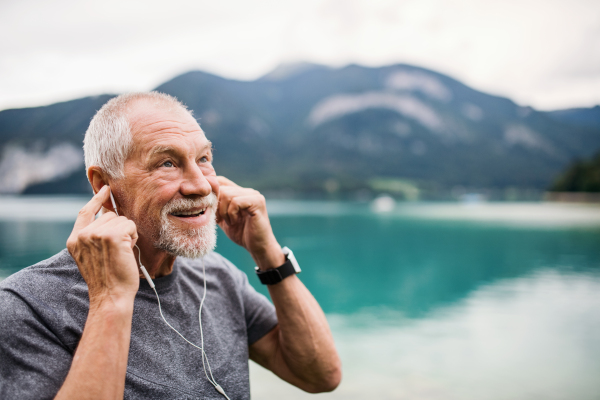 A senior man with earphones standing by lake in nature, listening to music.