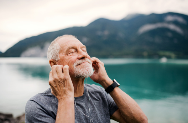 A senior man with earphones standing by lake in nature, listening to music.