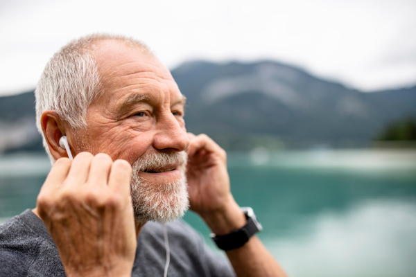 A senior man with earphones standing by lake in nature, listening to music.