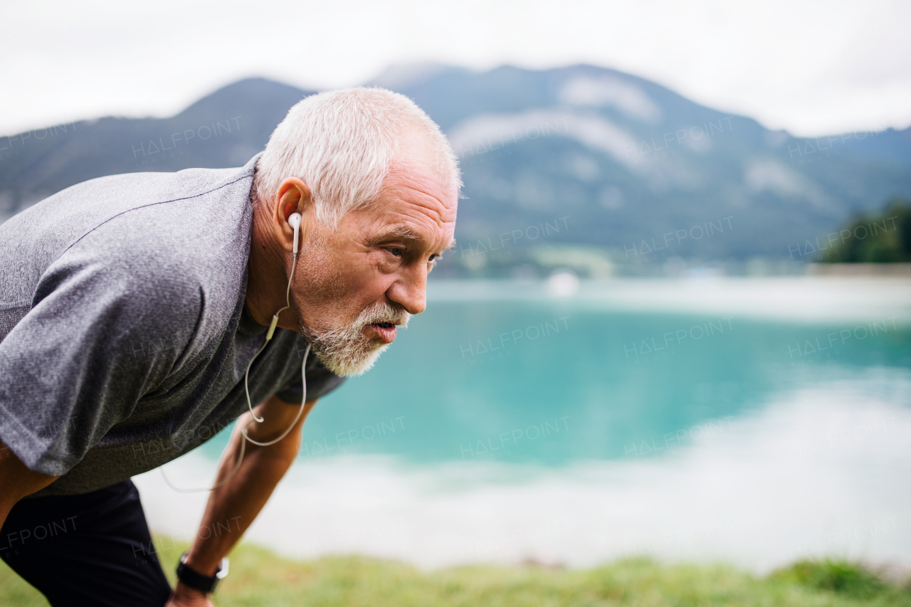 A senior man with earphones standing by lake in nature, doing exercise. Copy space.