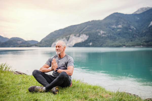 A senior man pensioner sitting by lake in nature, doing yoga exercise. Copy space.