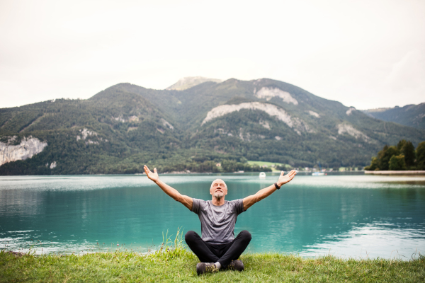 A senior man pensioner sitting by lake in nature, doing yoga exercise. Copy space.