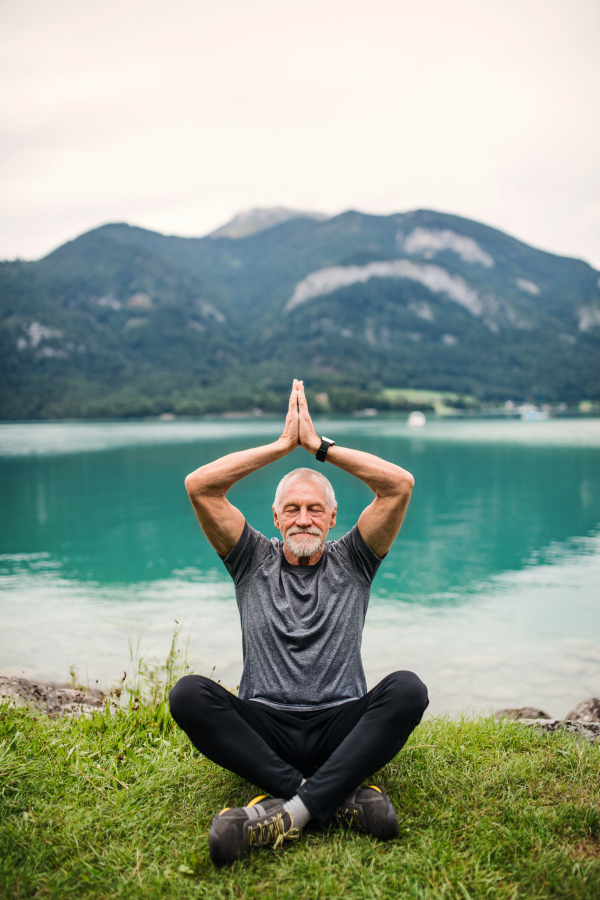 A front view of senior man pensioner sitting by lake in nature, doing yoga exercise.