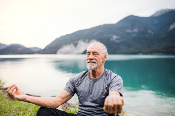 A senior man pensioner sitting by lake in nature, doing yoga exercise. Copy space.