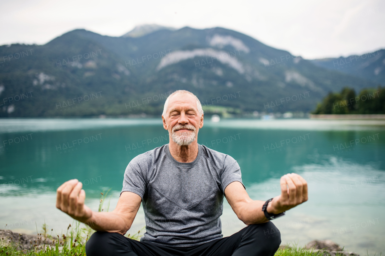 A senior man pensioner sitting by lake in nature, doing yoga exercise. Copy space.