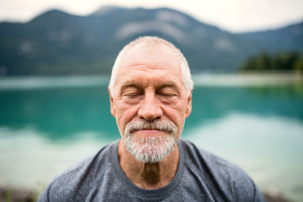 A front view portrait of senior man pensioner standing outdoors in nature, eyes closed.