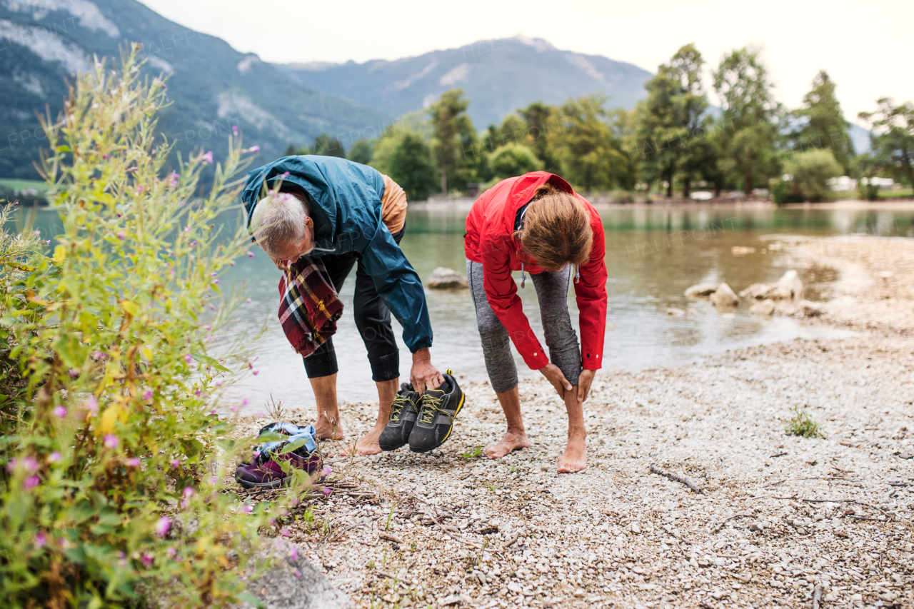 A front view of senior pensioner couple hikers standing by lake in nature, taking shoes off.