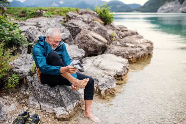 A senior man hiker sitting by lake in nature, taking shoes off. Copy space.