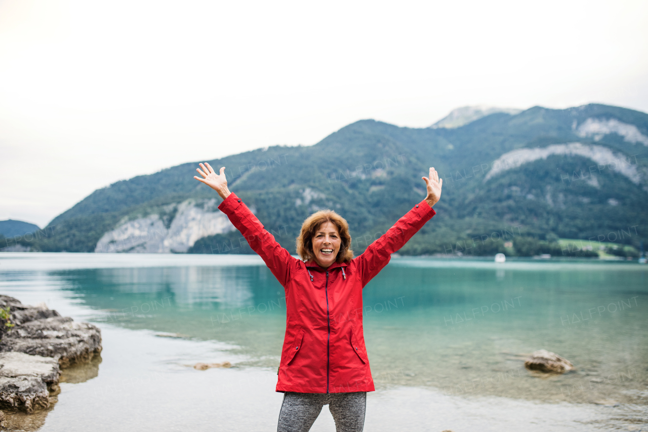 A front view of senior woman hiker standing by lake in nature, stretching arms.