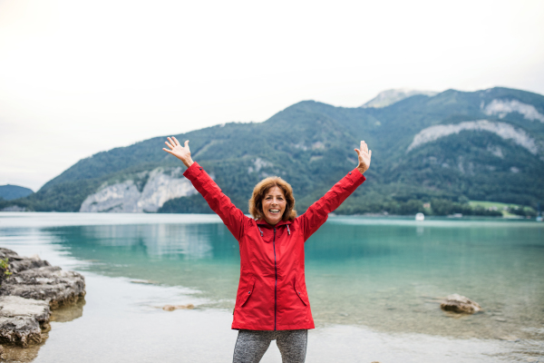 A front view of senior woman hiker standing by lake in nature, stretching arms.