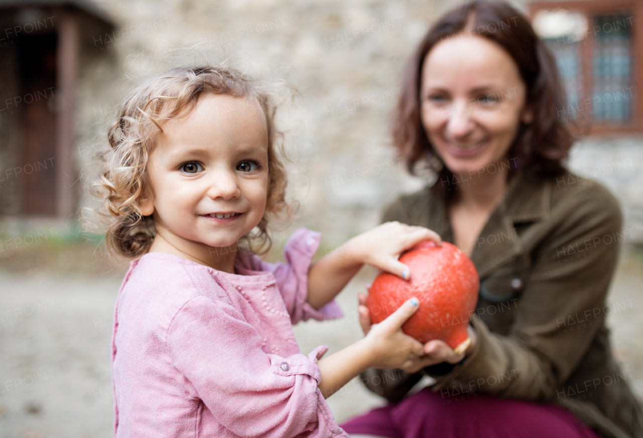 A small girl with mother standing in front of old stone house, holding hokkaido pumpkin.