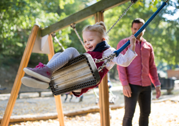 A cheerful small toddler girl with a father on a swing on a playground.