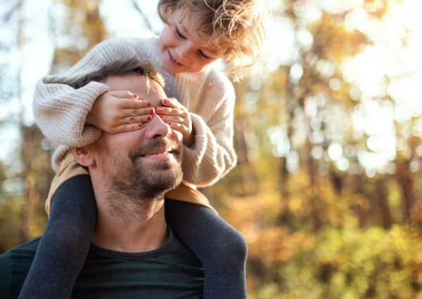 Mature father giving piggyback ride to happy small daughter on a walk in autumn forest.