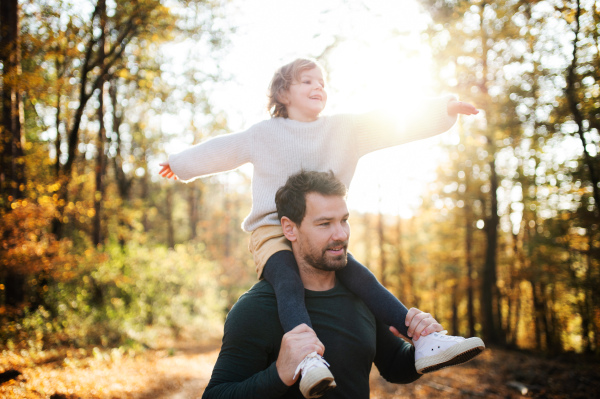 Mature father giving piggyback ride to happy small daughter on a walk in autumn forest.