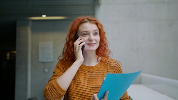 A young female university student walking through corridor indoors, having a call.