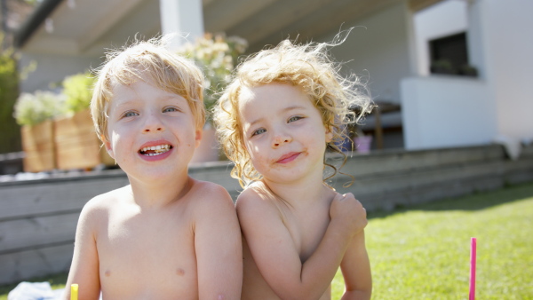 A front view of two sibling in swimsuit sitting next to pool and looking at camera.