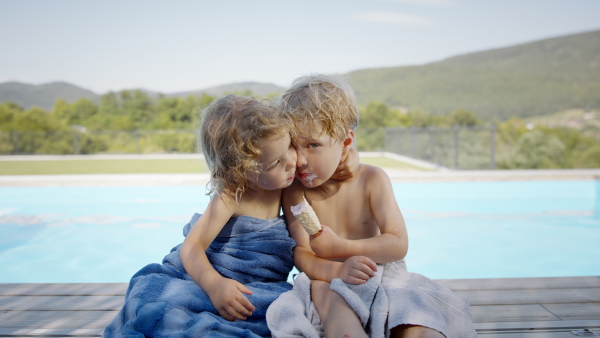 A front view of two sibling in swimsuit sitting next to pool, eating ice cream and looking at camera.