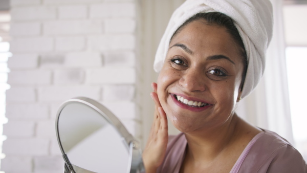 A close up of beautiful young overweight woman indoors at home, applying face cream and looking at camera.