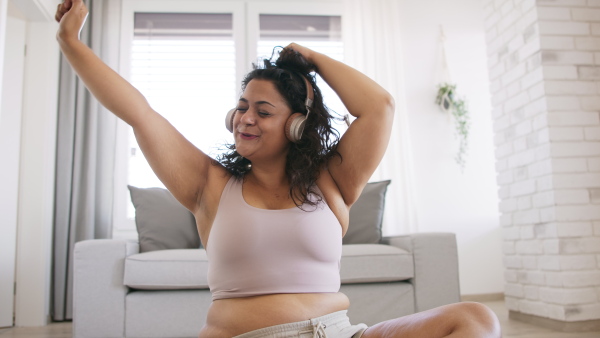 A happy beautiful young overweight woman sitting on floor indoors at home, listening music and dancing