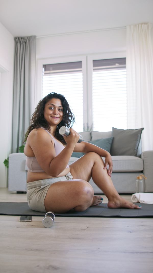 A vertical footage of happy young overweight woman indoors at home, exercising and looking at camera.