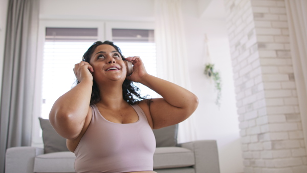 A happy beautiful young overweight woman sitting on floor indoors at home, listening music and dancing