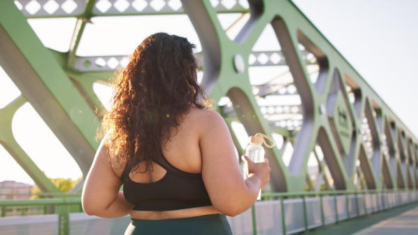 A rear view of young overweight woman in sports clothes walking outdoors in bridge.