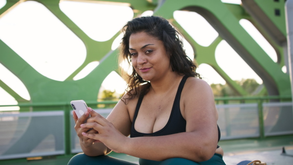 A young overweight woman in sports clothes sitting and using smartphone outdoors in bridge.