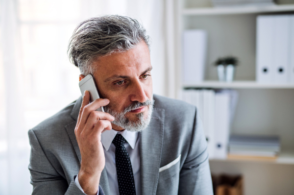 A serious mature businessman with smartphone sitting at the table in an office, making a phone call.