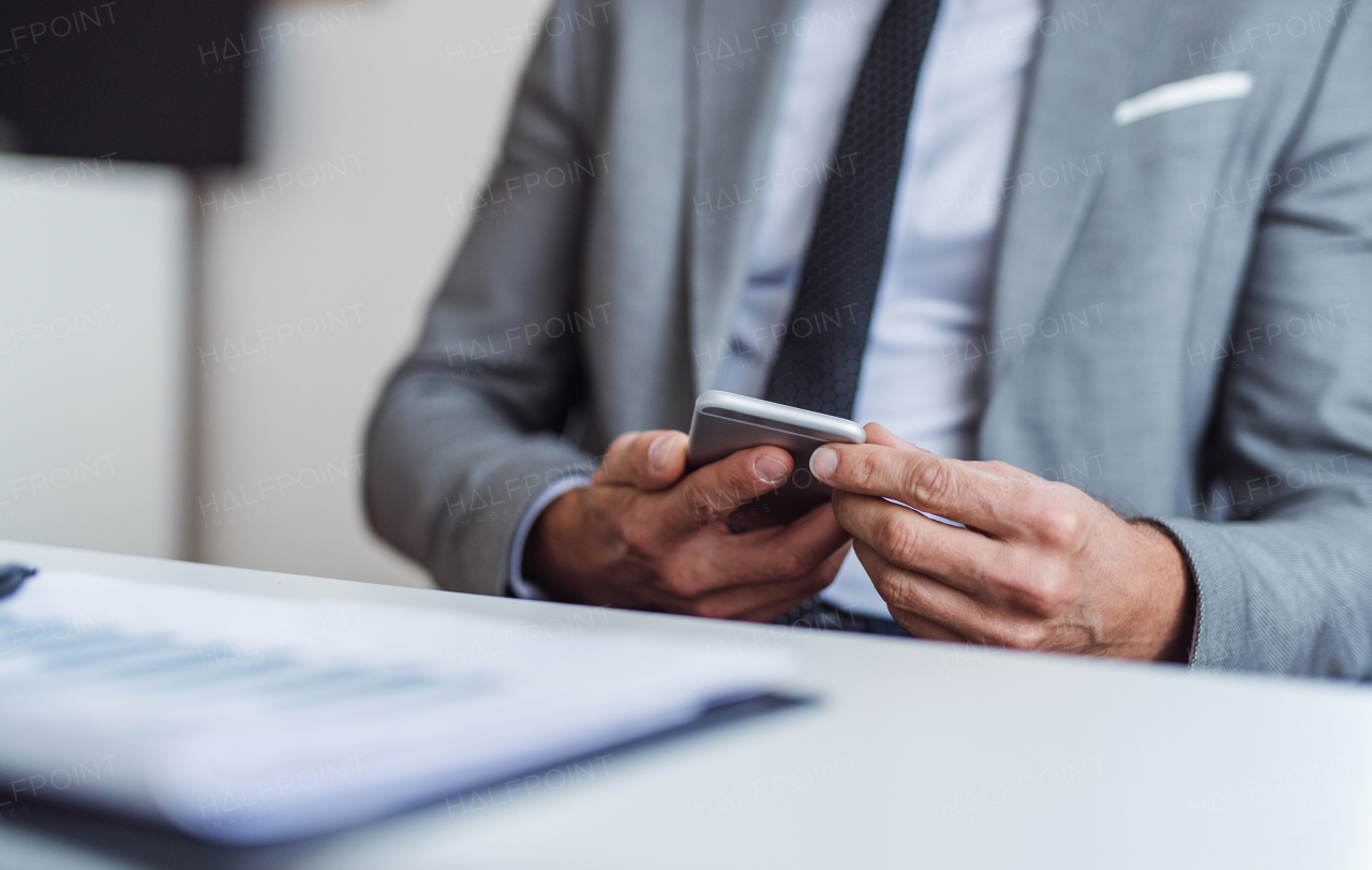 A midsection of businessman with smartphone and laptop sitting at the table in an office, working. Copy space.