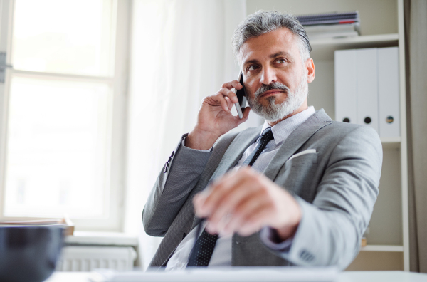 A serious mature businessman with smartphone sitting at the table in an office, making a phone call.